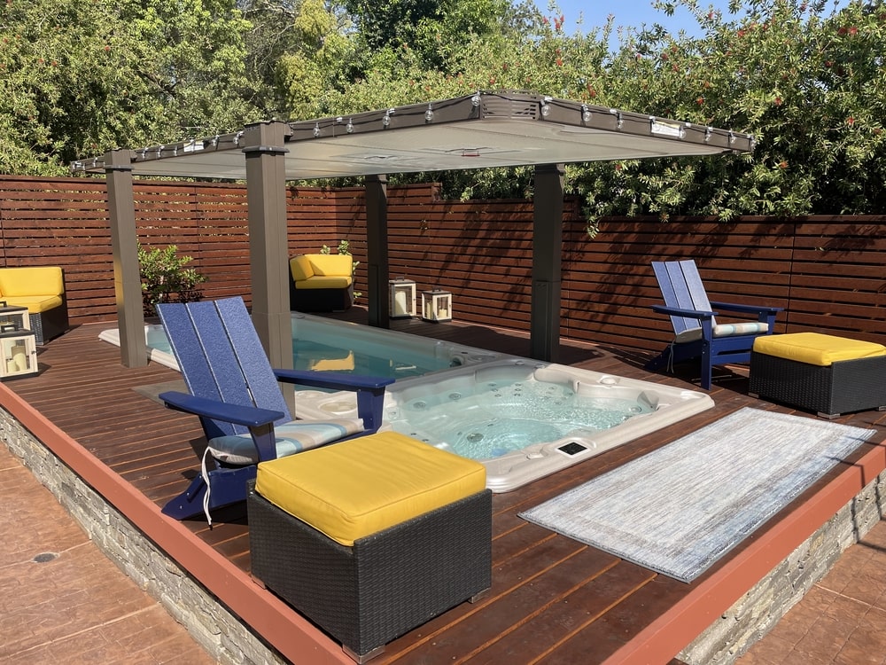 A hot tub in a backyard with a pergola and blue lounge chairs