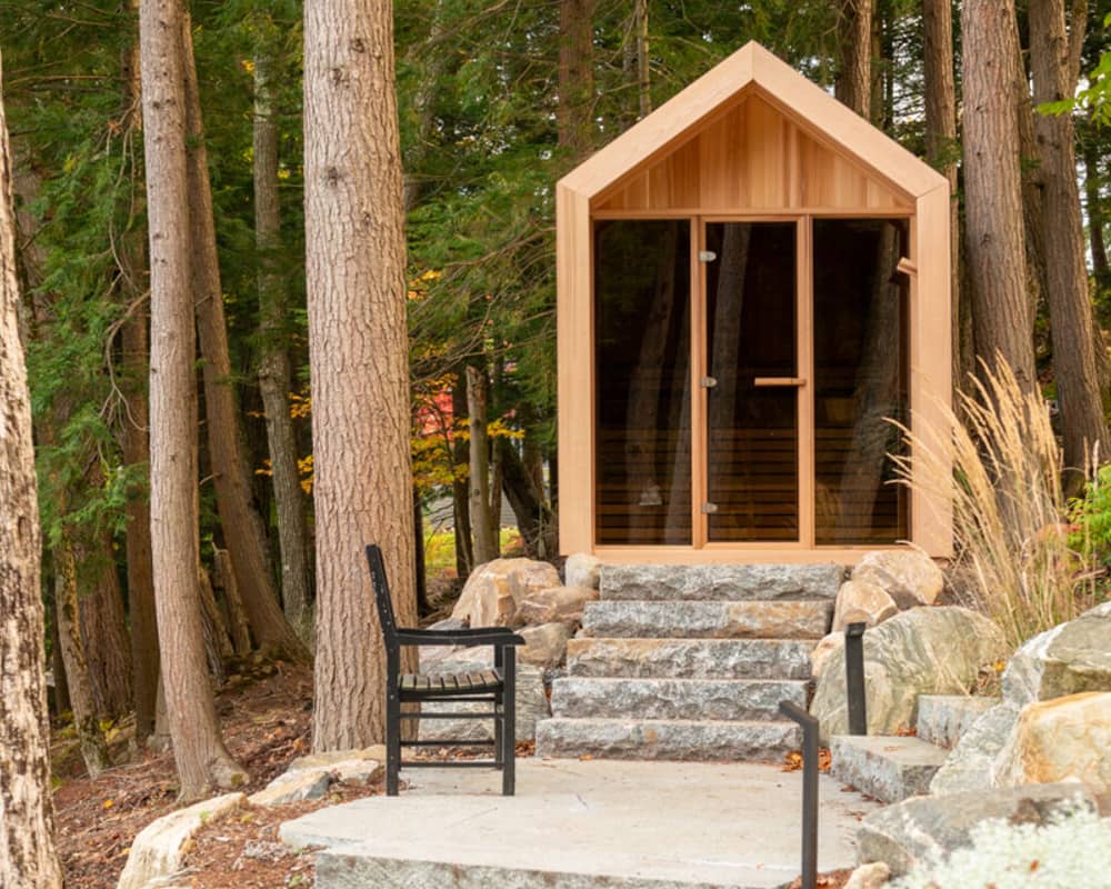 Leisurecraft Pure Cube Hudson Sauna  with glass doors in a forested area with a black chair and stone steps.