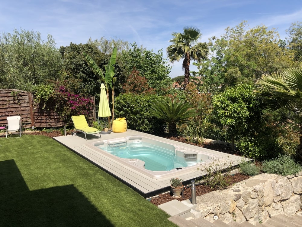 A backyard with a swim spa and a yellow umbrella||father is playing with kids in swim spa|A hot tub in a backyard with a pergola and blue lounge chairs